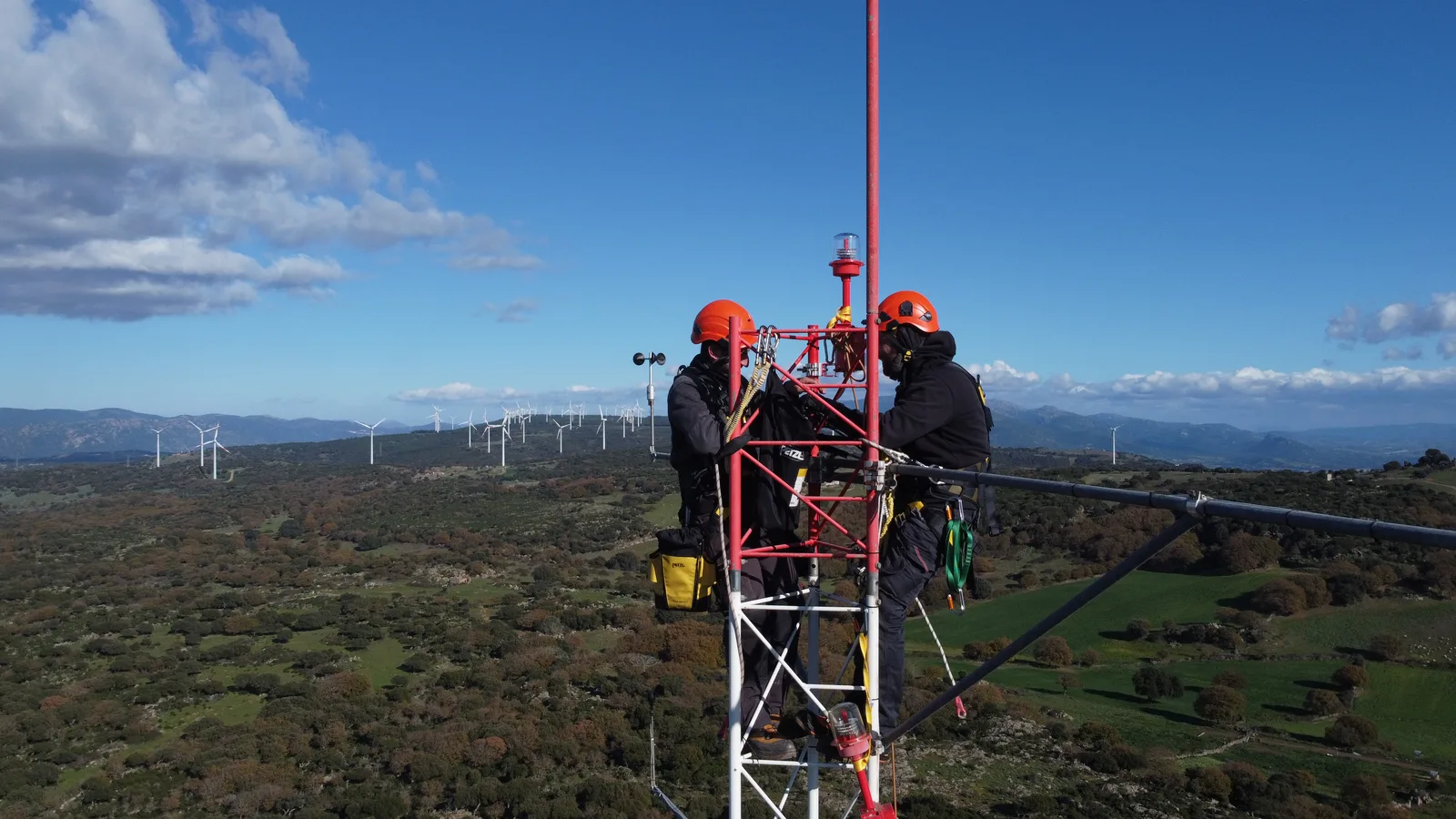 Due tecnici Tecnogaia con imbracatura e casco lavorano in cima a un traliccio anemometrico, con un parco eolico all'orizzonte