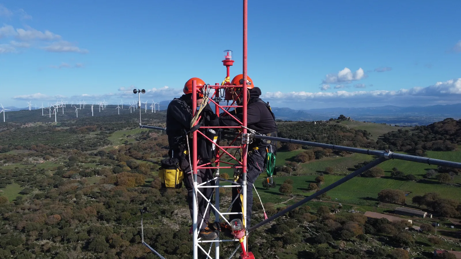 Tecnici al lavoro sui sensori in cima a un traliccio, campagna e aerogeneratori sullo sfondo