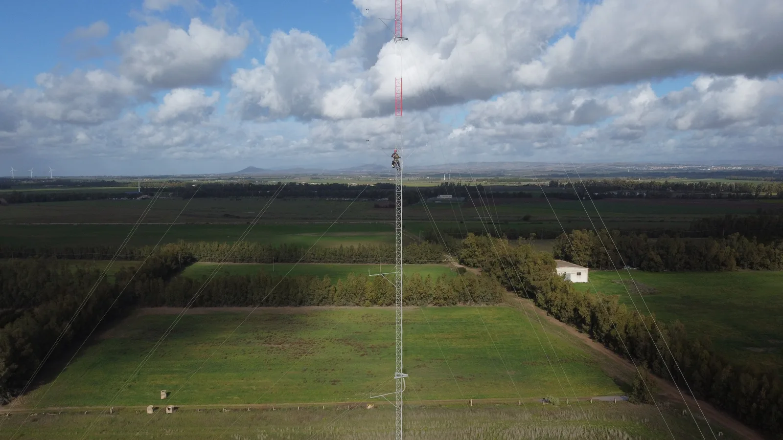 Ripresa aerea di un traliccio anemometrico che si alza sopra i campi, cielo con nuvole